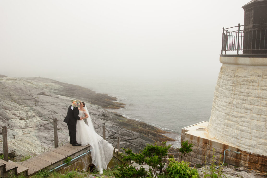 Bride and groom kissing on the rocky coastline beside a lighthouse in Newport Rhode Island