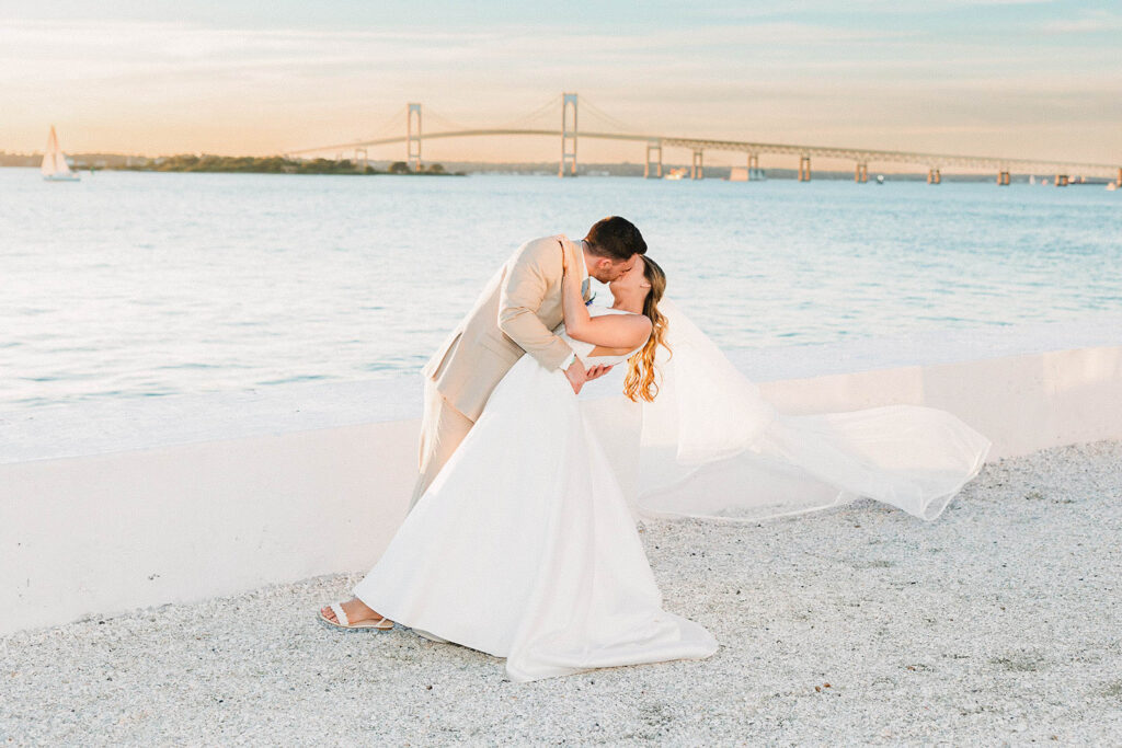 Allison and Christian embracing along the Belle Mer seawall with Narragansett Bay and the Newport Bridge beyond
