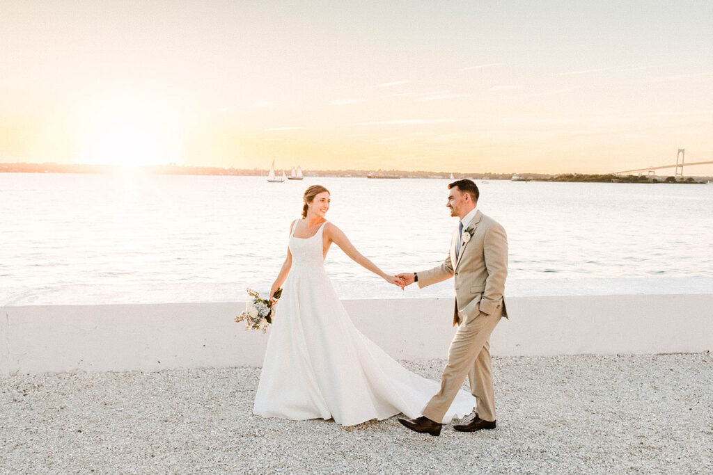 Allison and Christian walking hand in hand along Narragansett Bay at Belle Mer with the Newport Bridge in the distance

